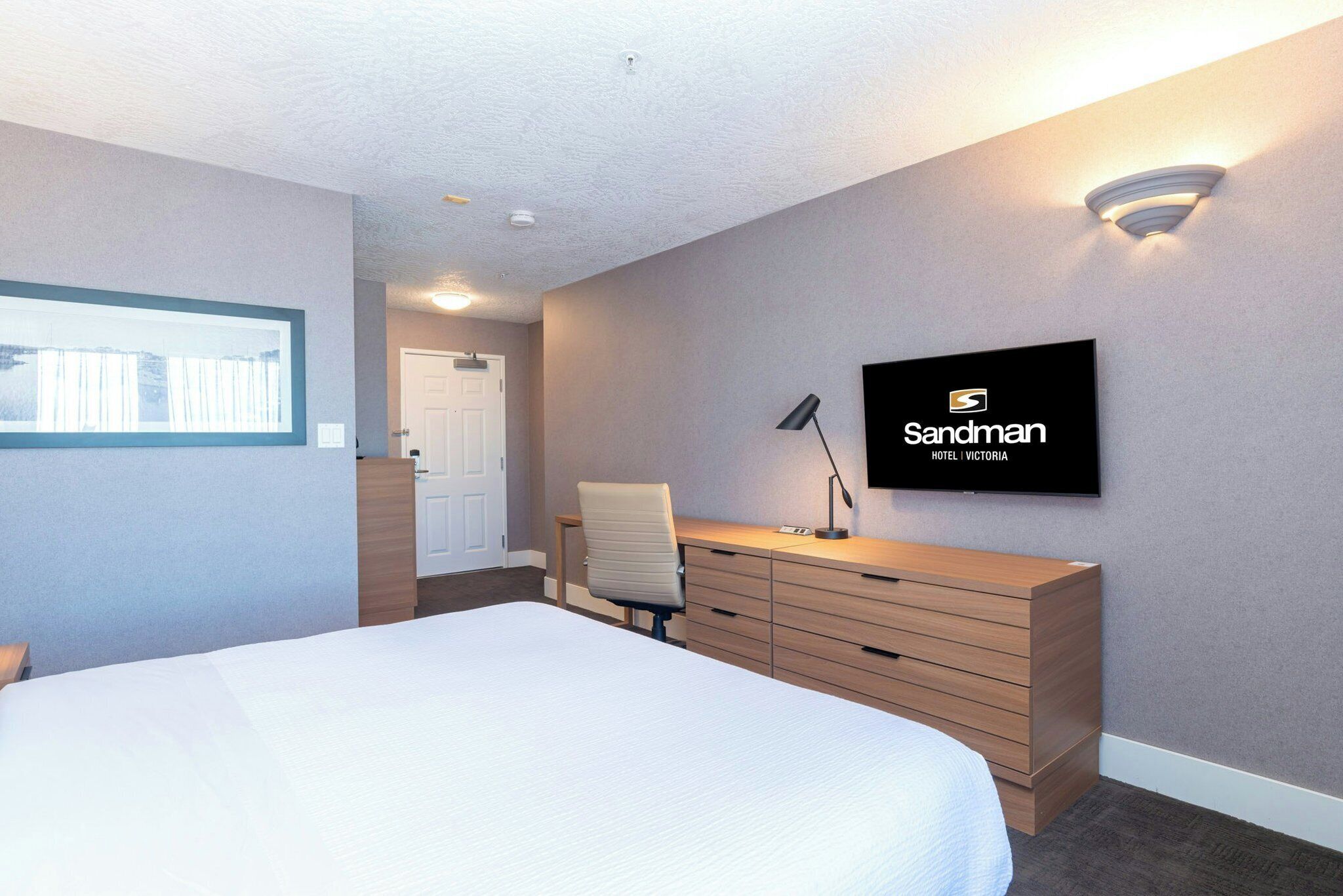 Une chambre d’hôtel comprenant un lit, un bureau avec des tiroirs, une chaise, un téléviseur fixé au mur et une porte donnant sur une autre pièce.