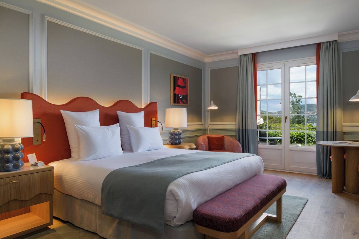 A hotel room with a large bed, two bedside tables, a bench, a window with curtains, and a view of greenery.