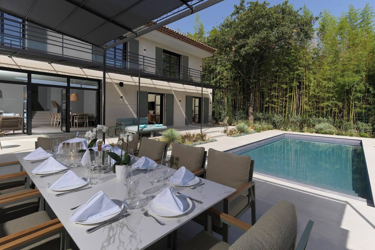 A dining area with a table set for four, outdoor pool, and a modern house with a balcony.