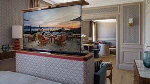 A hotel room with a large flat-screen TV mounted on a wooden console, a bed, a seating area with a sofa and chairs, and a view of the outdoors through a large window. - Althoff Hotel Villa Belrose (Gassin)