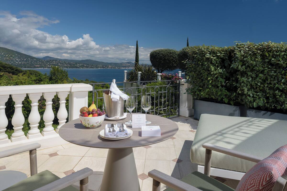 A round table with a white bowl of fruit, a pitcher, and wine glasses on a balcony overlooking a coastal landscape.