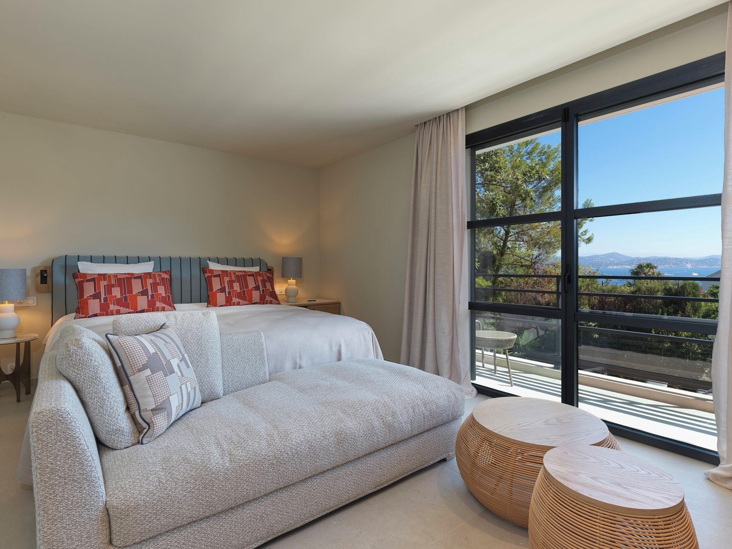 A modern hotel room with a large bed, a sofa, a small table, and a view of the sea.