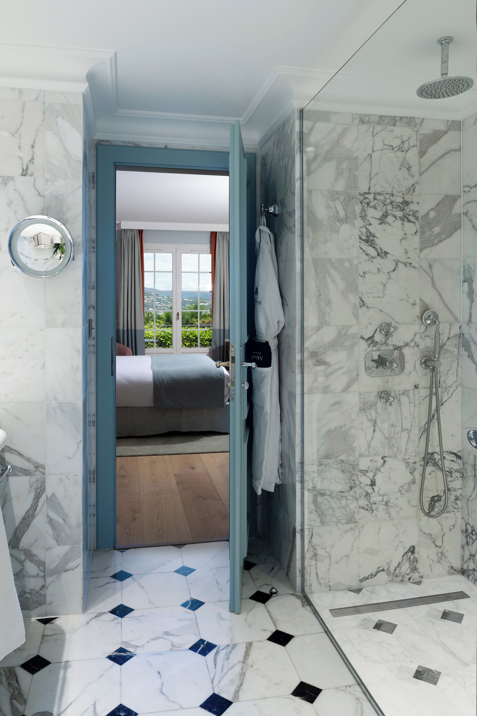 Ein Badezimmer mit Dusche, Toilette und Waschbecken. Eine Tür führt zu einem Schlafzimmer mit Bett und Blick ins Grüne.