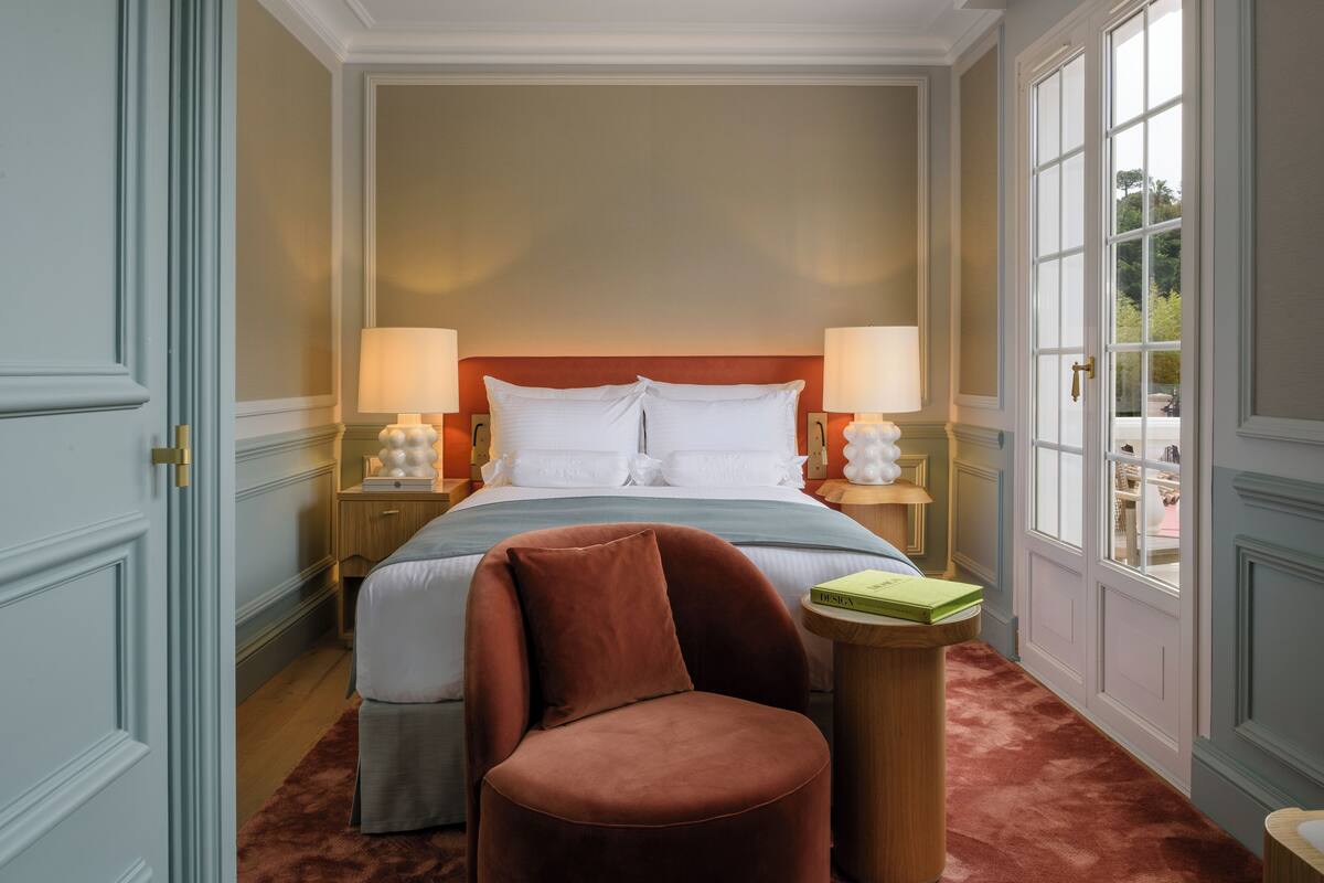 A hotel room with a large bed, two bedside lamps, a brown armchair, a small wooden side table, and a view of the outdoors through a large window.