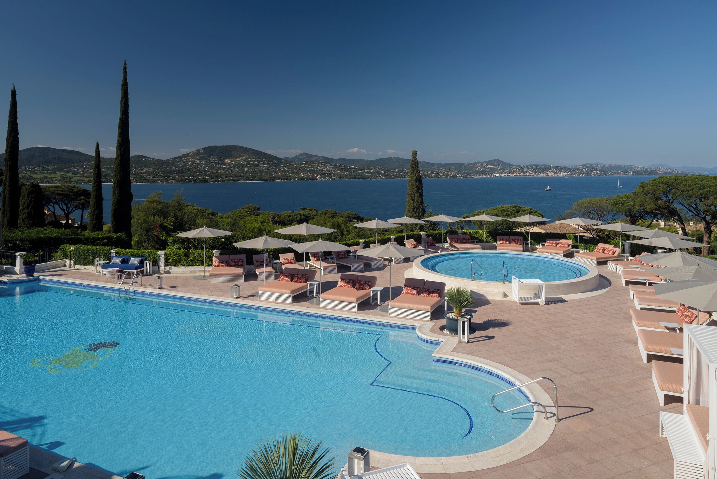 A pool area with lounge chairs and umbrellas, surrounded by trees and overlooking a body of water.