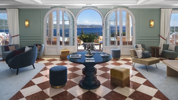 A spacious lounge with a checkered floor, a central round table, and seating arrangements. Large windows with glass doors provide a view of the sea. - Althoff Hotel Villa Belrose (Gassin)