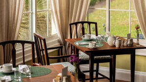 A dining area with a table set for two, chairs, a window with curtains, and a view of greenery outside. - Casa Velha do Palheiro, Relais & Chateaux (Funchal)