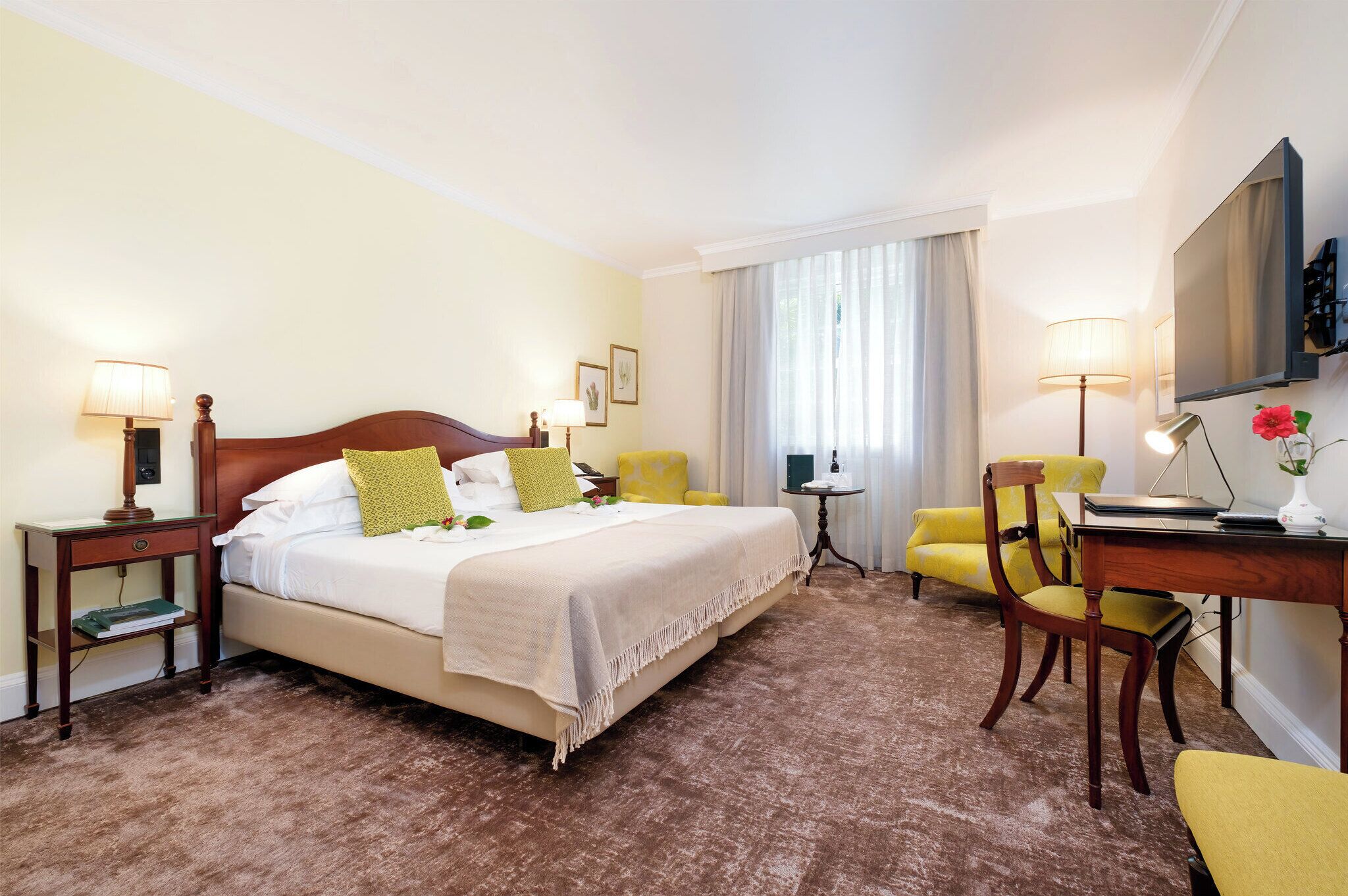 A hotel room with a bed, two chairs, a desk, a TV, and a window with curtains.
