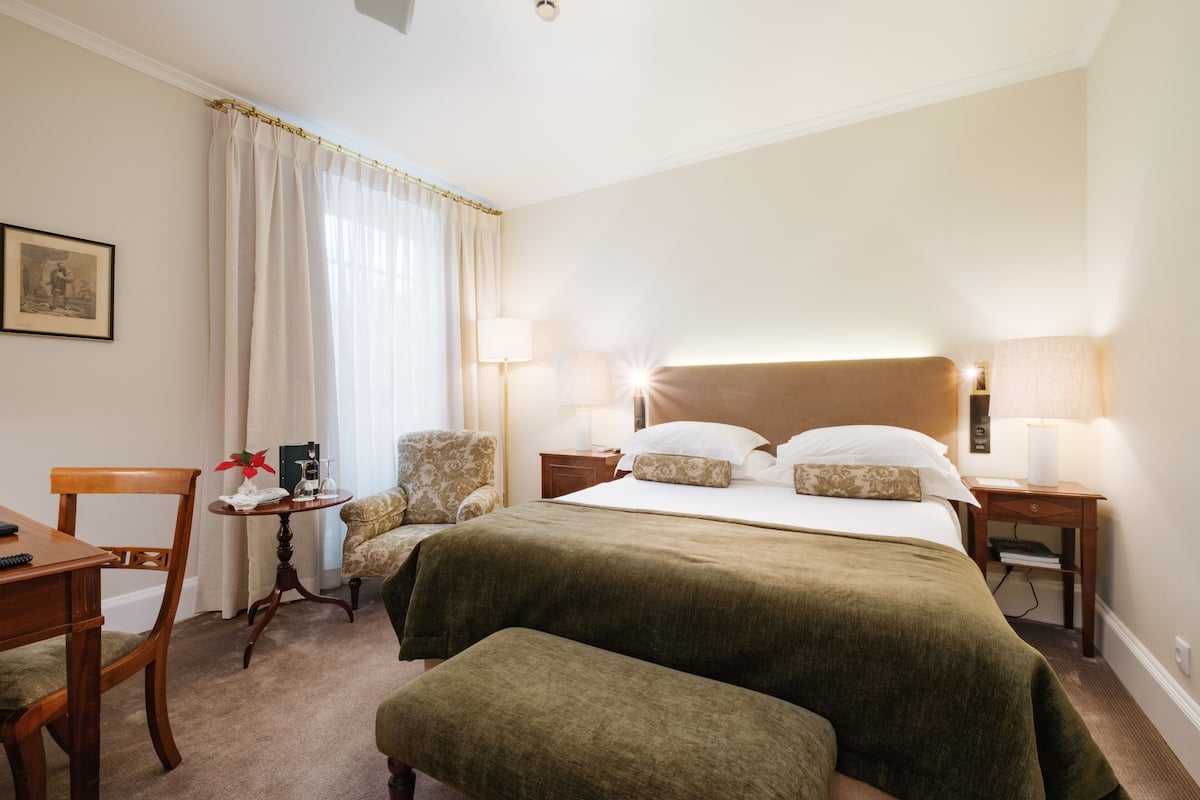 Una habitación de hotel con una cama grande, dos mesitas de noche con lámparas, una silla, una pequeña mesa con una planta y un cuadro en la pared.