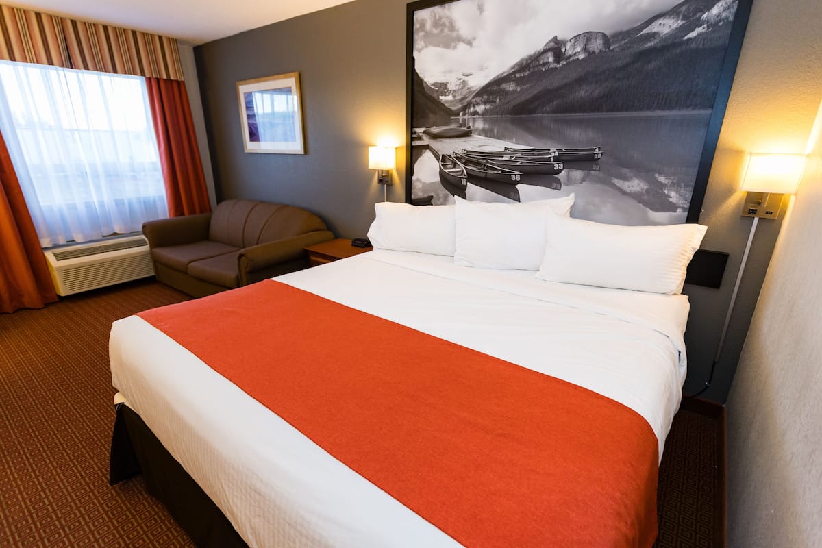A hotel room with a large bed, a sofa, a window with curtains, a wall-mounted lamp, and a framed picture of a mountain landscape.