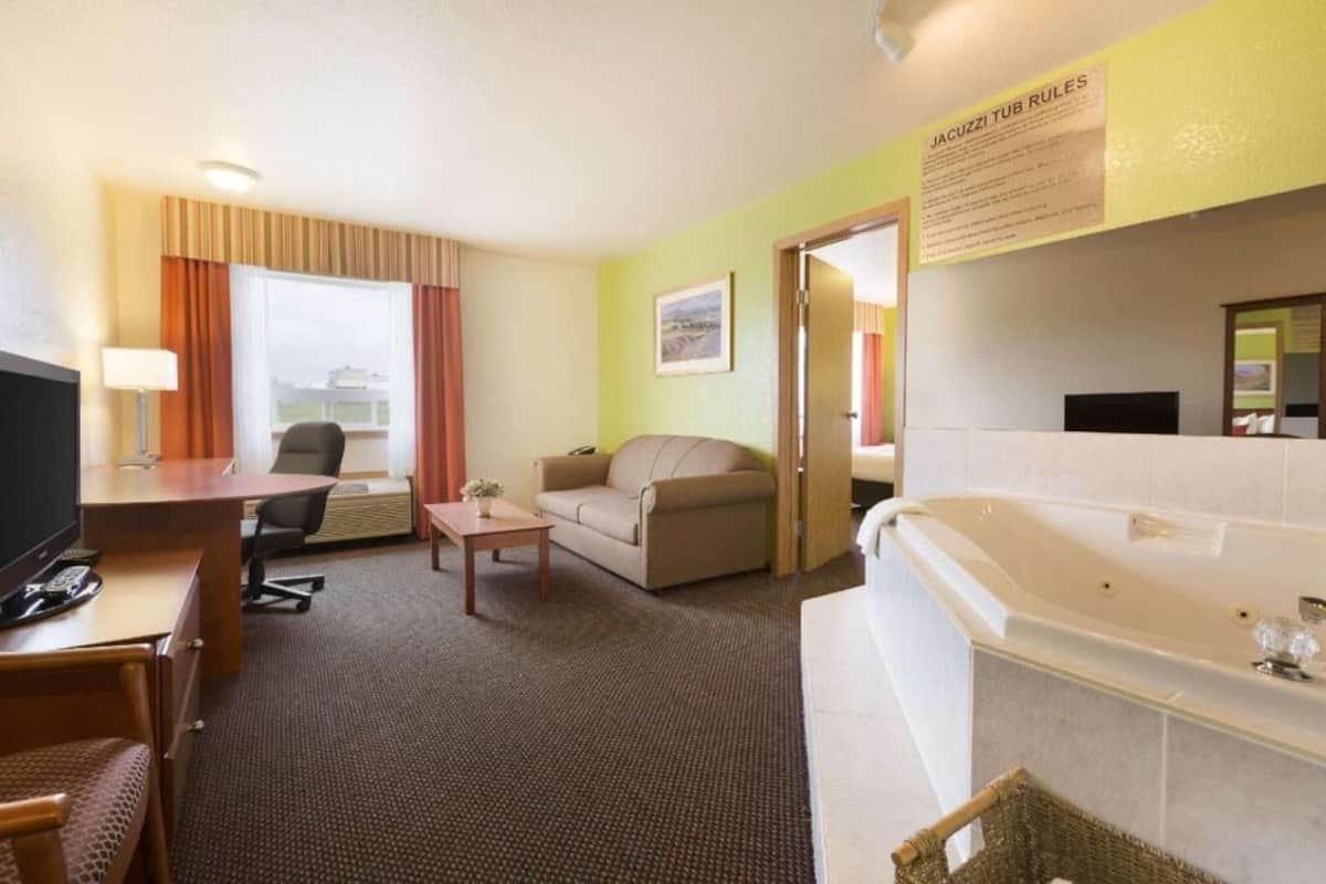 A hotel room with a desk, a sofa, a small table, a TV, a bathtub, and a window with curtains.