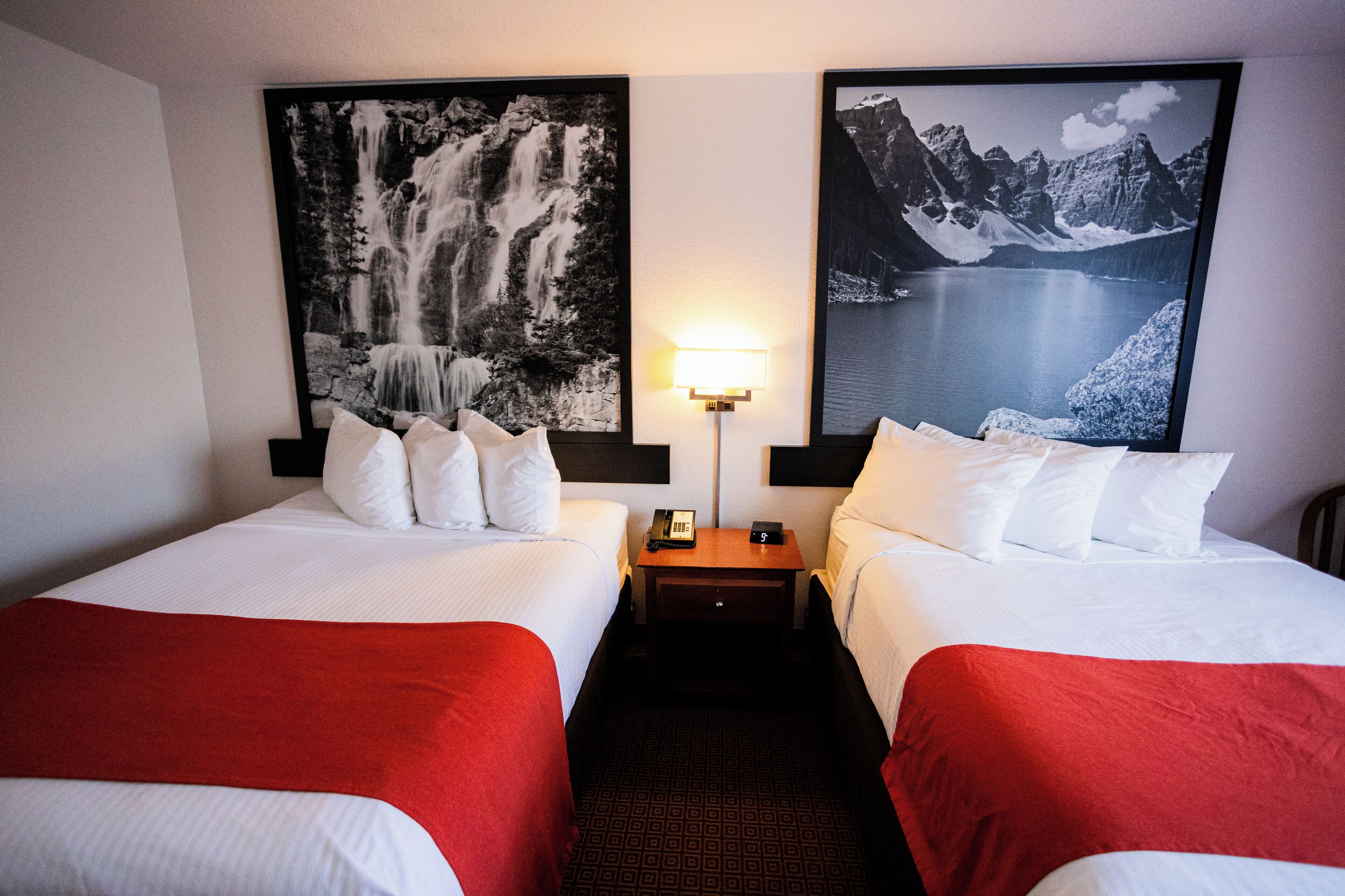 A hotel room with two beds, each with white bedding and red throw blankets. There are two large framed pictures on the walls, one of a waterfall and the other of a mountain lake. A bedside table with a lamp and a phone is between the beds.