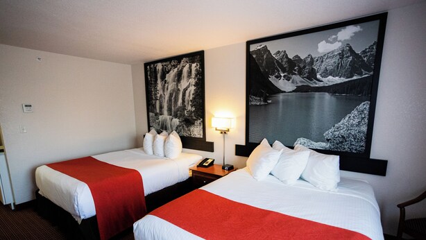 A hotel room with two beds, each with white bedding and red throw blankets. There are two wall-mounted paintings, one of a waterfall and the other of a mountain lake. A bedside table with a lamp is visible between the beds. - Super 8 by Wyndham Drayton Valley (Drayton Valley)