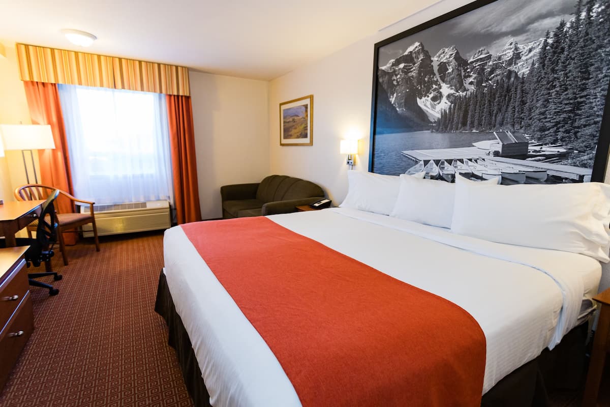 A hotel room with a large bed, a desk with a chair, a desk lamp, and a painting of a mountainous landscape.