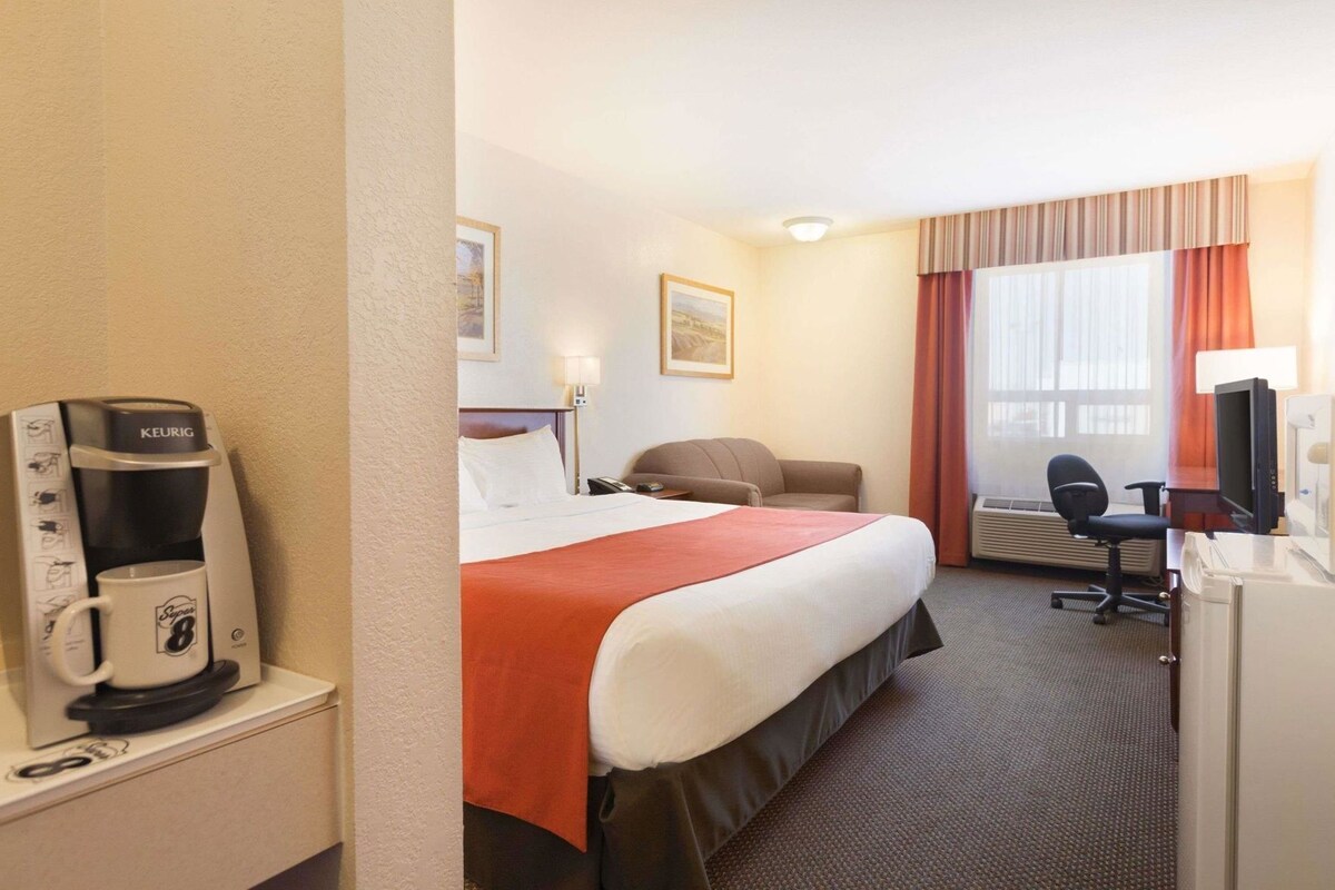 Habitación de hotel con cama, escritorio, silla, televisión, cafetera y ventana con cortinas.