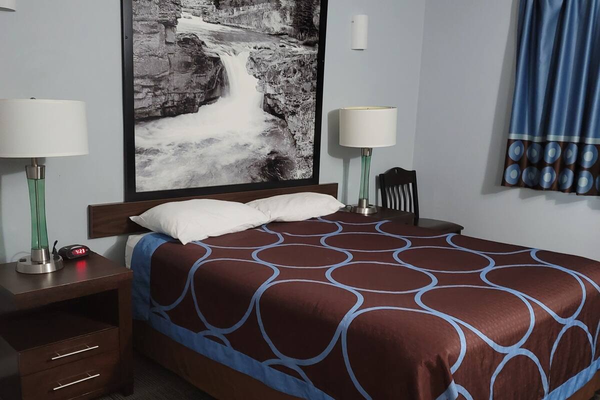 A hotel room with a large bed, two bedside tables with lamps, a clock, a framed picture on the wall, and a window with curtains.