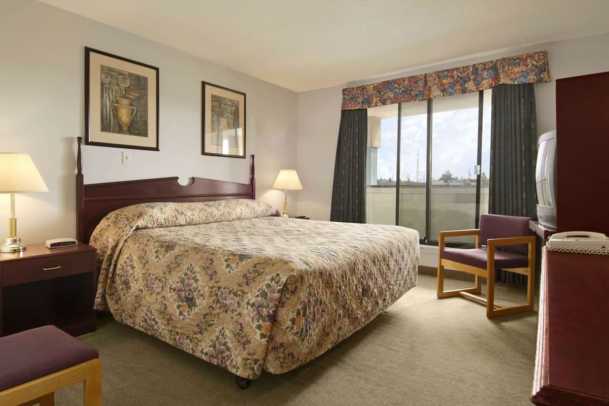 A hotel room with a large bed, two bedside tables with lamps, a chair, a television, and a window with curtains.