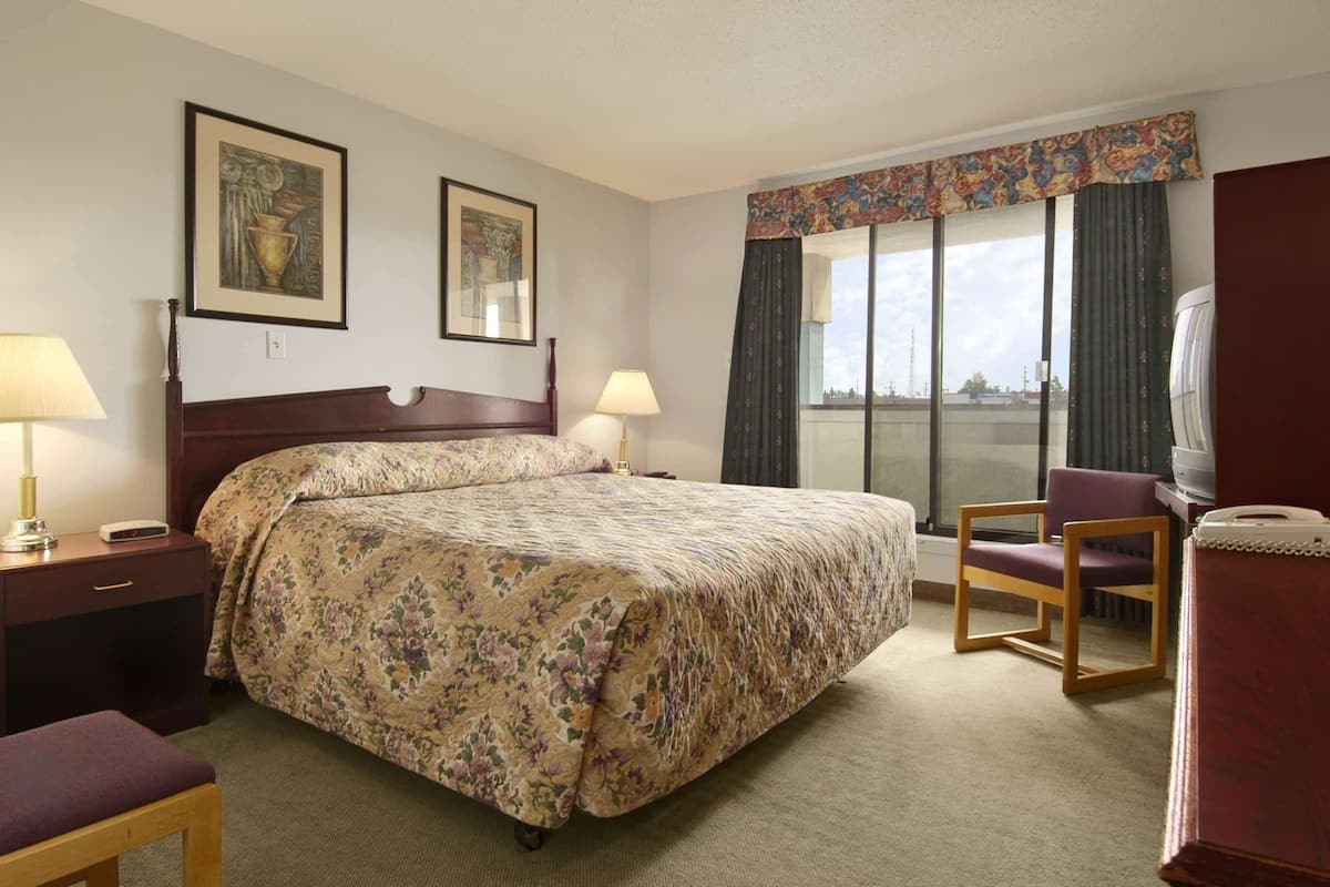 A hotel room with a large bed, two bedside tables with lamps, a chair, a TV, and a window with curtains.
