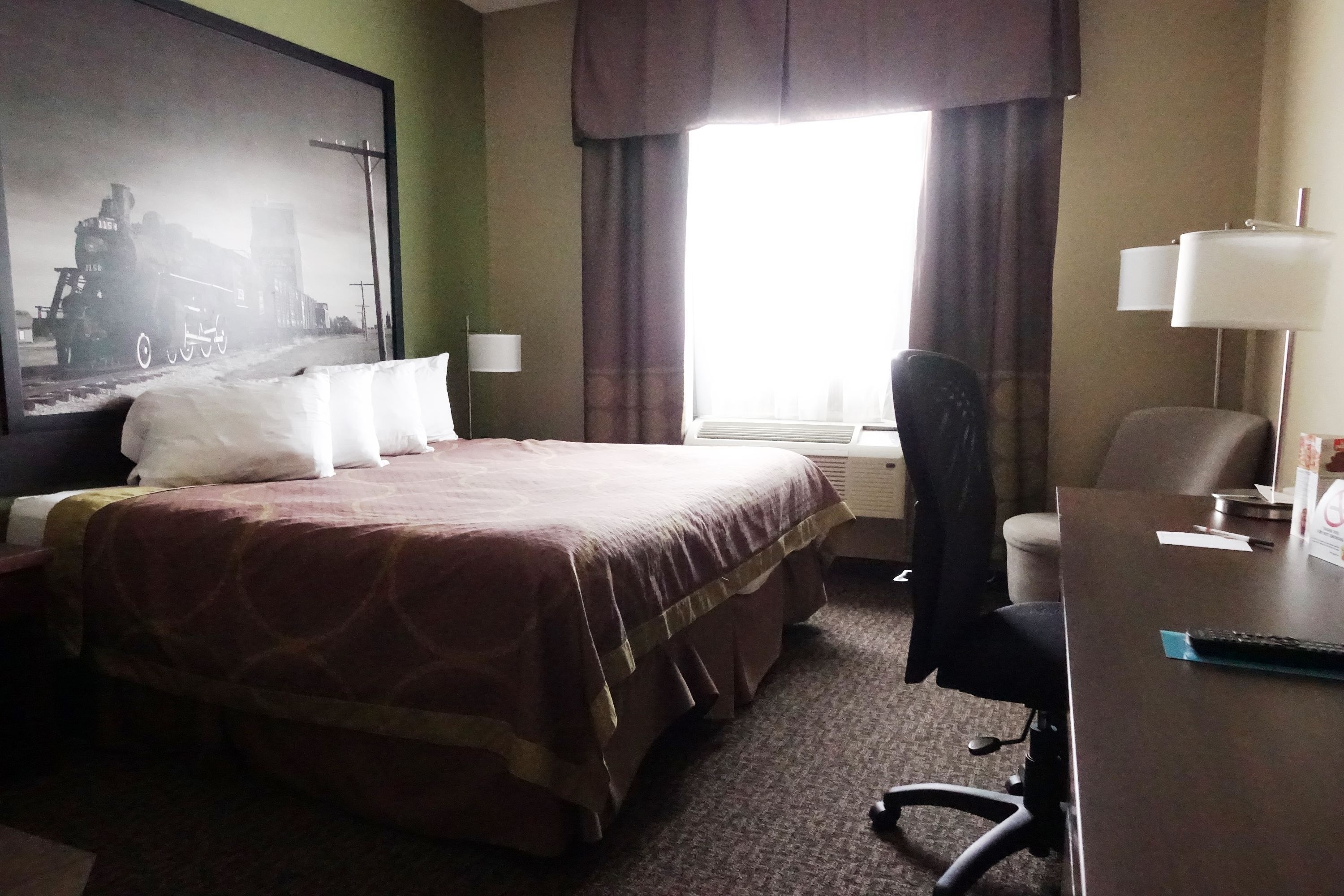 Una habitación de hotel con una cama grande, un escritorio, una silla, una lámpara y un ventanal con cortinas.
