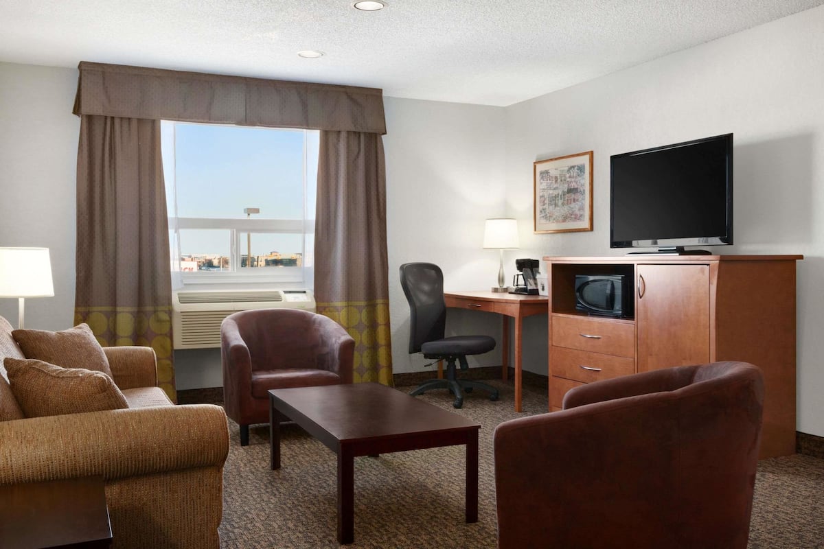 Une chambre d’hôtel avec un canapé, deux fauteuils, un bureau avec un ordinateur, une télévision et une fenêtre avec des rideaux.
