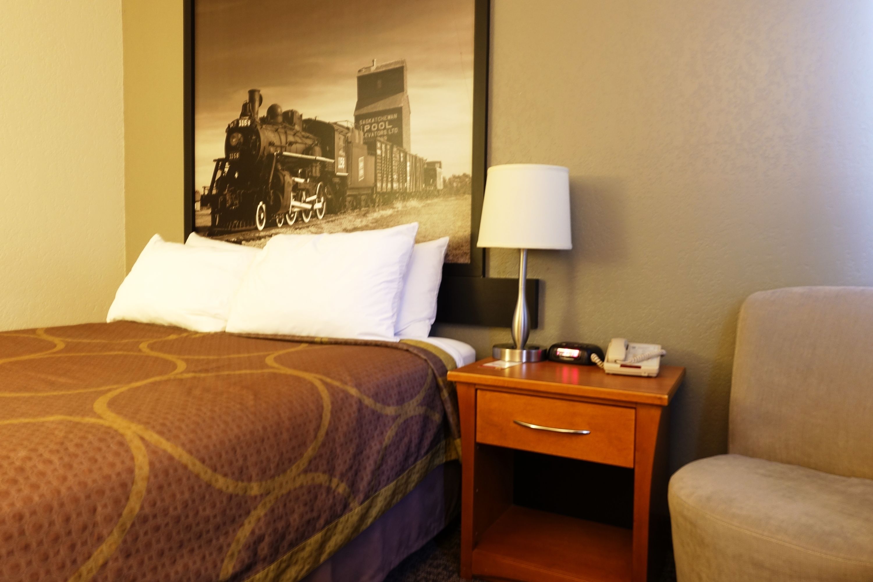 A hotel room with a bed, a nightstand with a lamp, a chair, and a large framed picture on the wall.