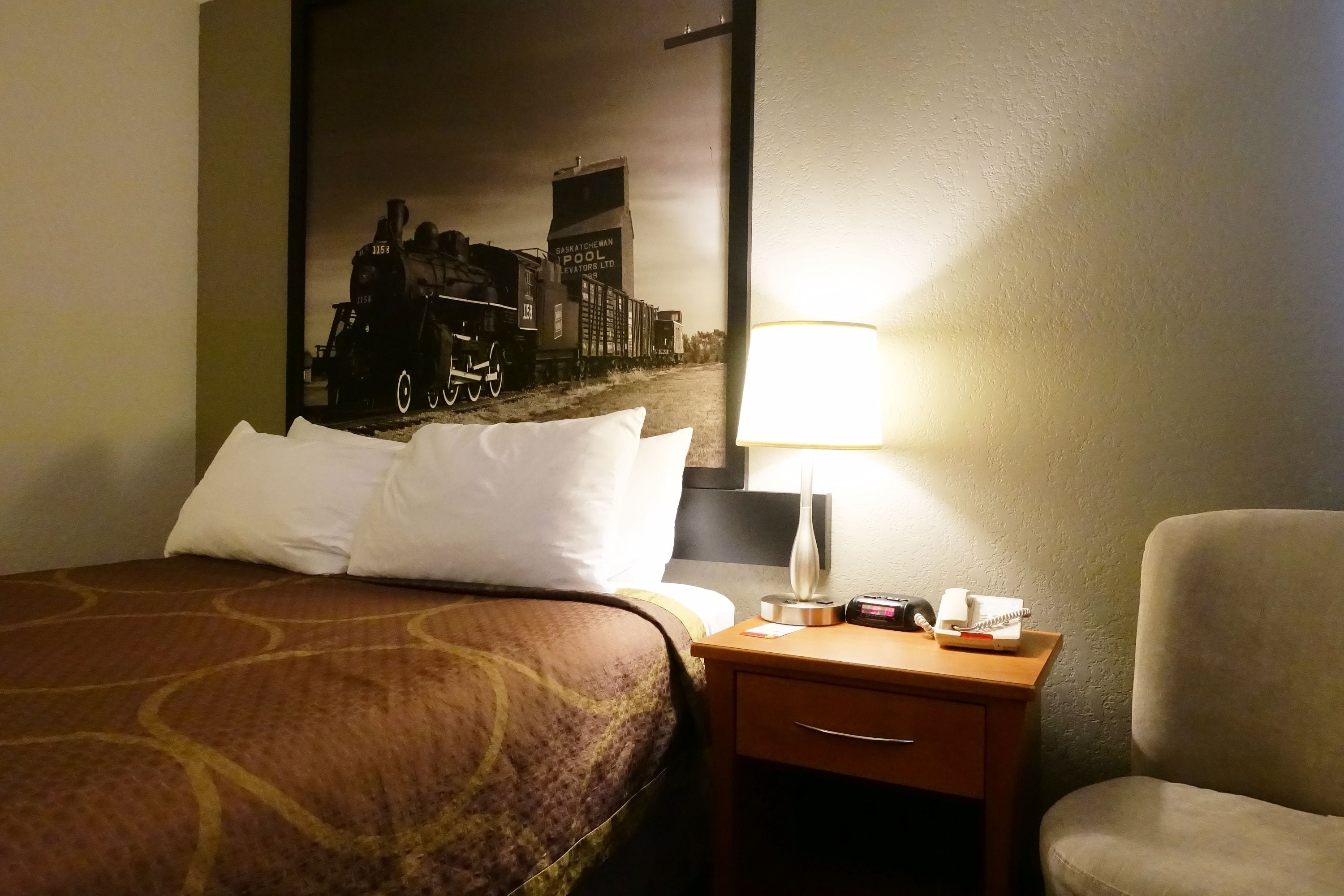 Ein Hotelzimmer mit einem Bett, einem Nachttisch mit Lampe, einem Sessel und einem an der Wand hängenden Bild von einem Zug.
