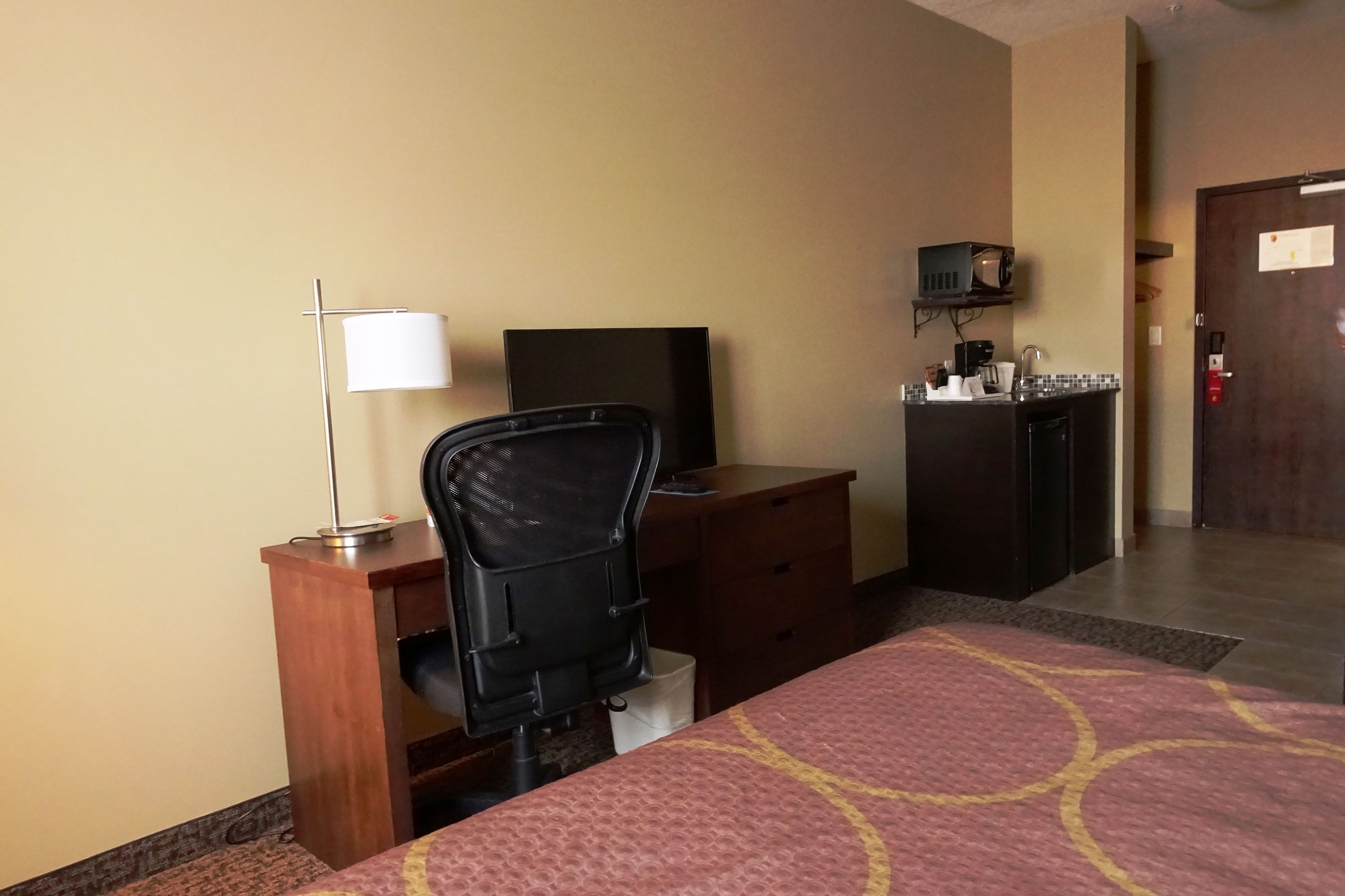 A hotel room with a bed, desk, chair, television, and a small kitchenette area.