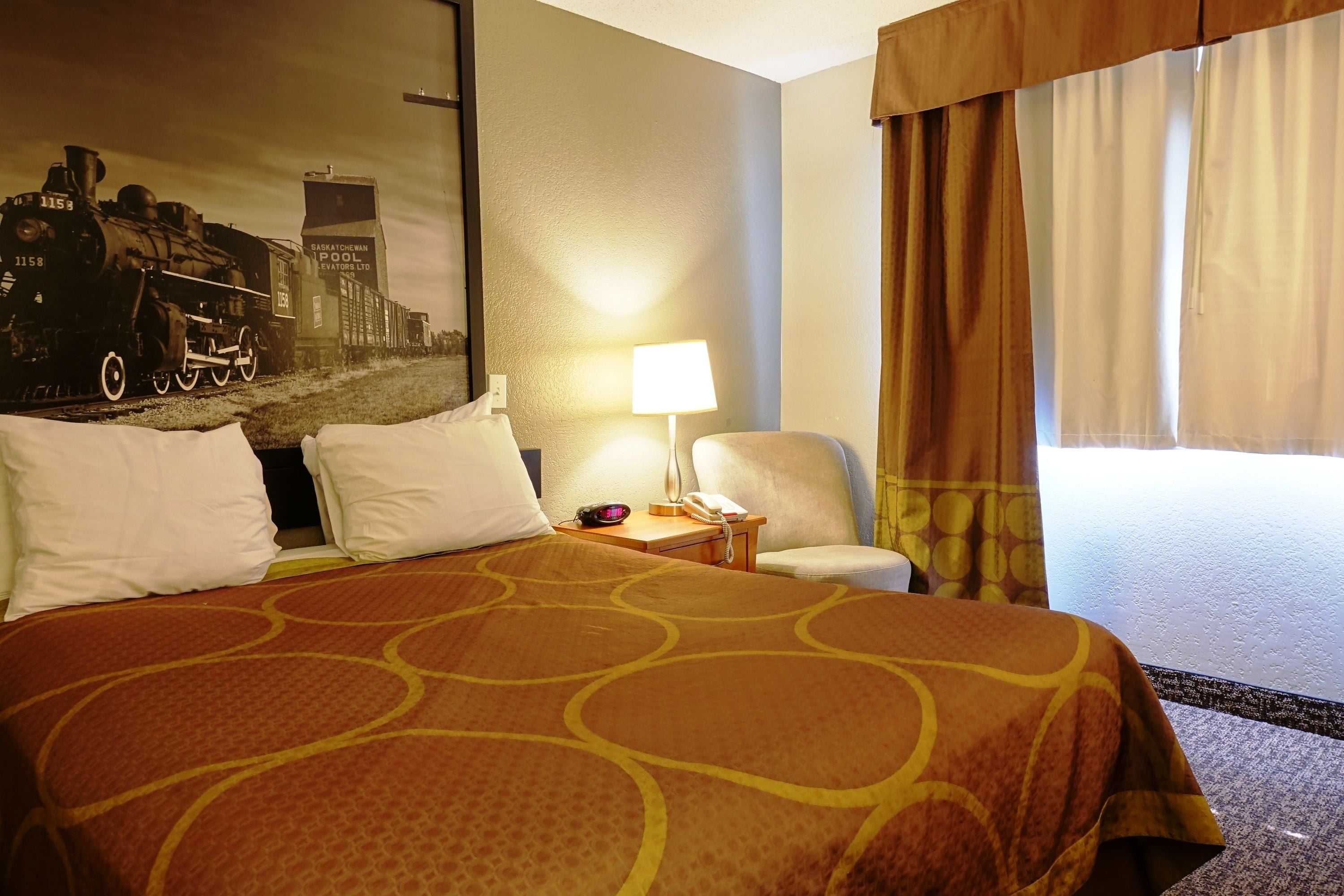 A hotel room with a large bed, a desk with a lamp, a chair, and a painting of a train on the wall.