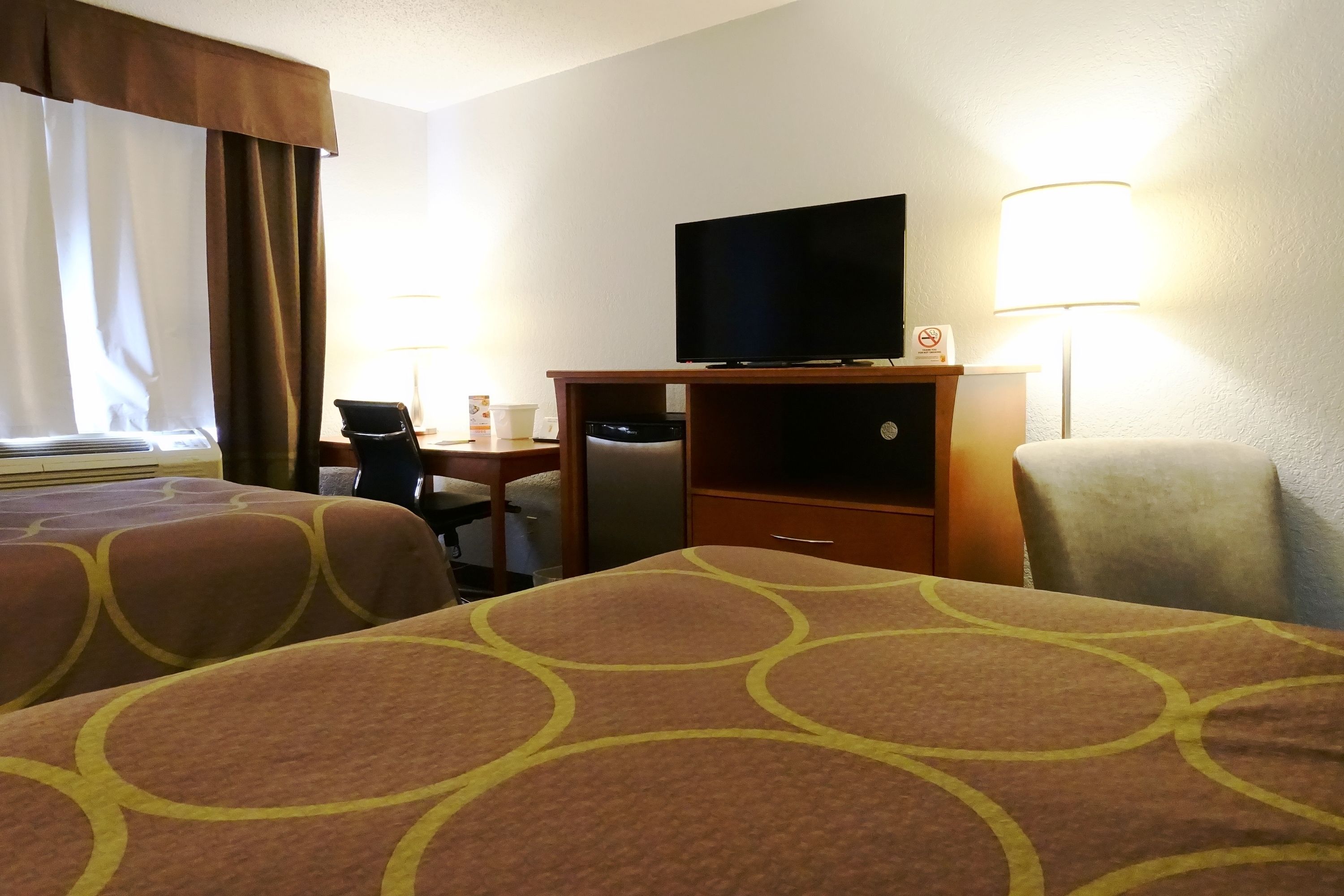 A hotel room with two beds, a desk with a television, a chair, a lamp, and a small table with a cup and a book.