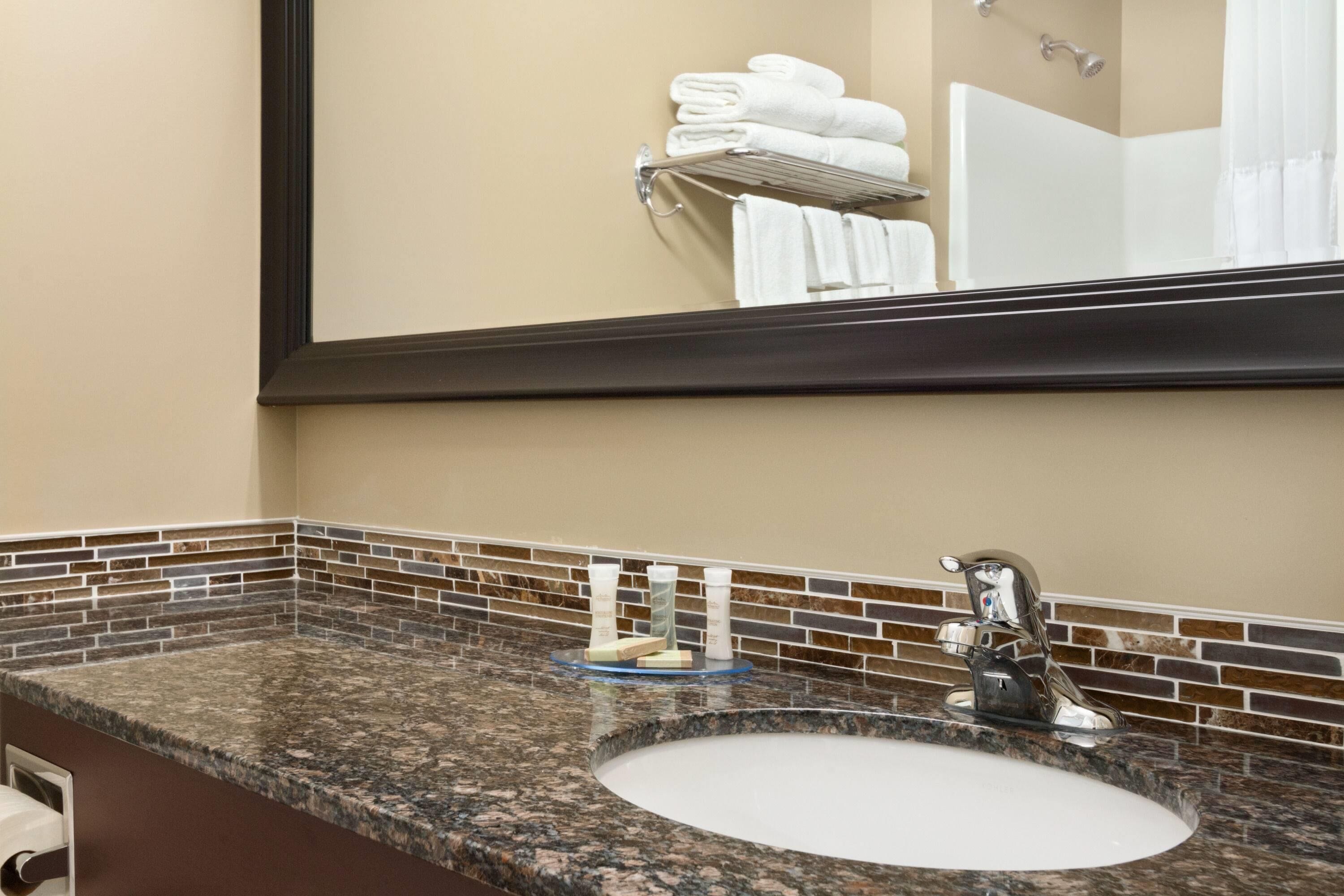 Baño con encimera de granito, lavamanos y espejo que refleja un perchero y la zona de la ducha.