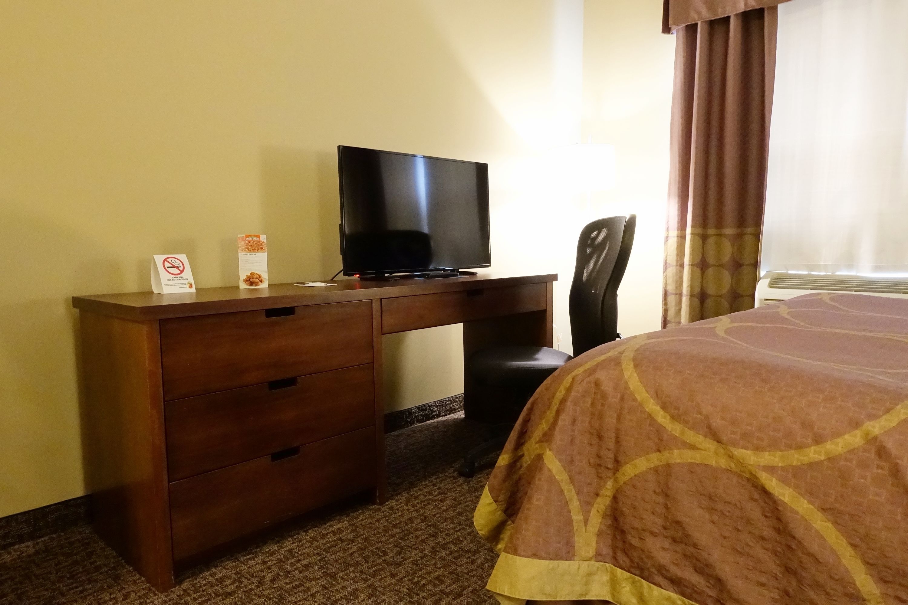 A hotel room with a wooden desk, a television, a chair, and a bed with a patterned bedspread.