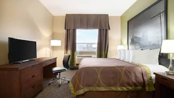 Quarto de hotel com cama, escrivaninha com televisão, cadeira, janela com cortinas e um quadro na parede.