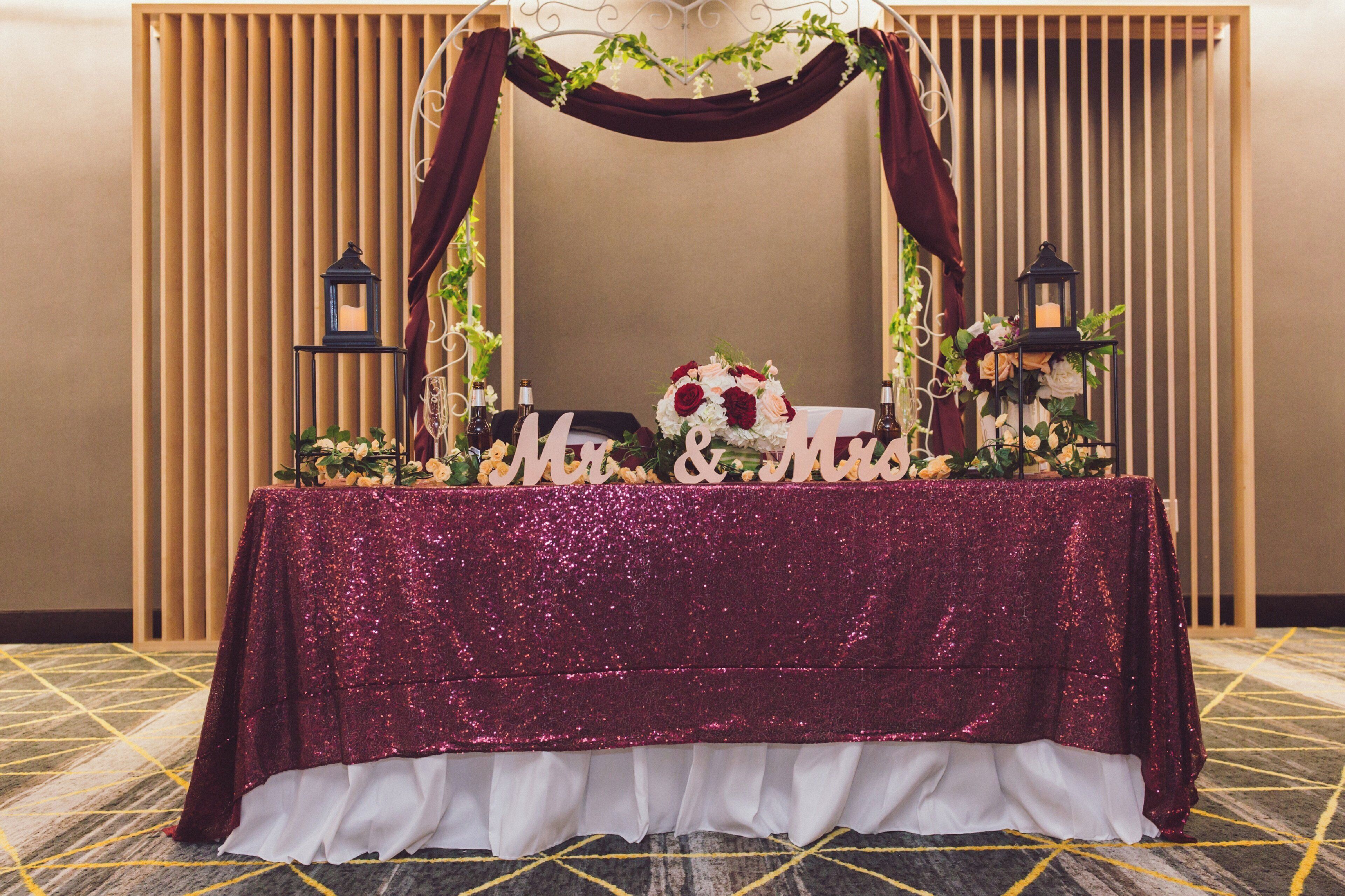 Una mesa con un mantel brillante, un arreglo floral y dos faroles.
