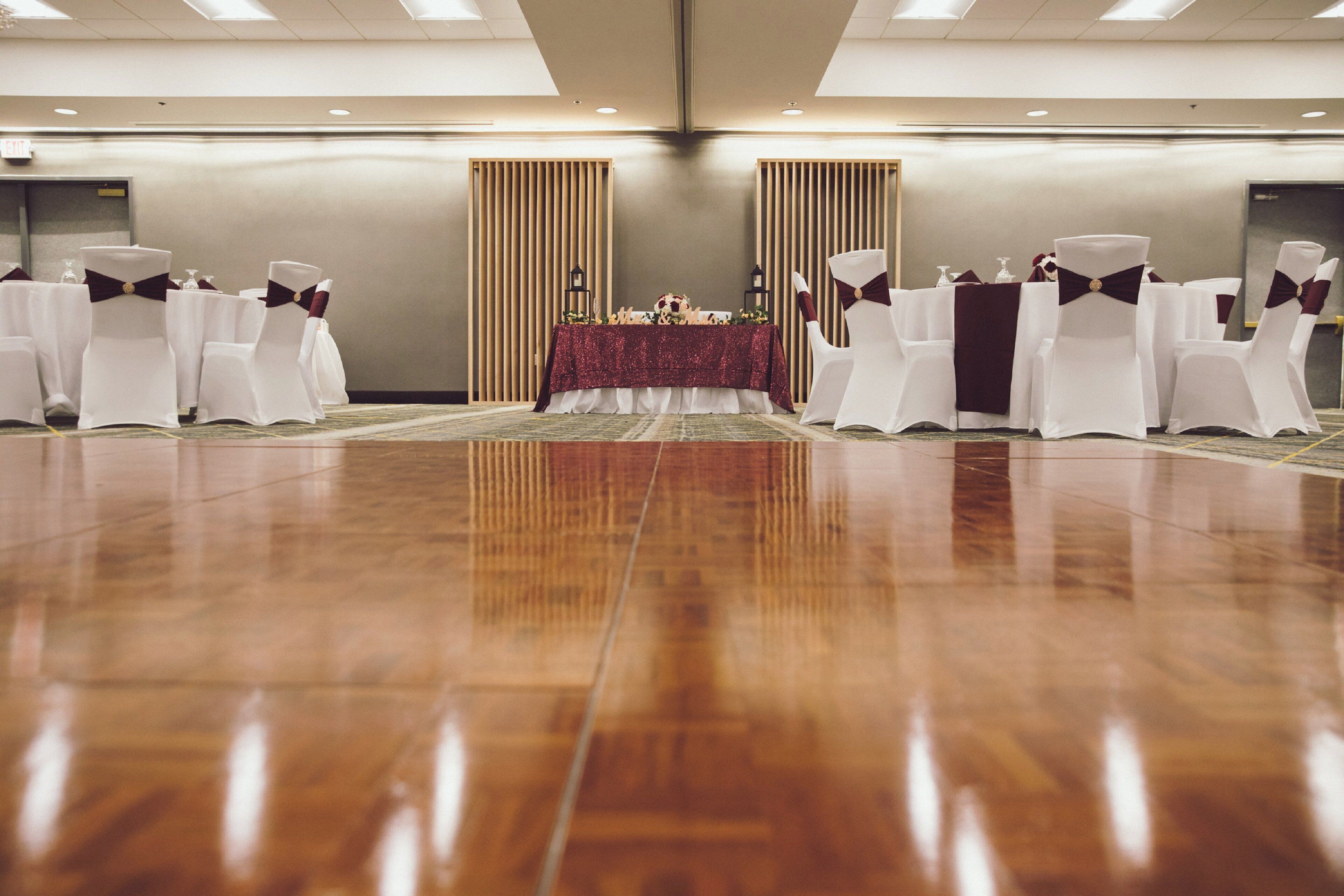 Un salón de conferencias con piso de madera pulida, sillas blancas con cintas oscuras y una mesa con mantel rojo.