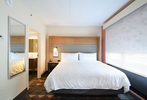 Een hotelkamer met een groot bed, een nachtkastje met lamp, een spiegel en uitzicht door een raam.