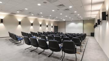 Un salón de conferencias con filas de sillas, una pantalla de proyector y luces en el techo.