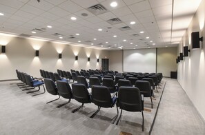 A conference room with rows of chairs, a projector screen, and ceiling lights. - Holiday Inn Fredericksburg Conference Ctr by IHG (Fredericksburg)