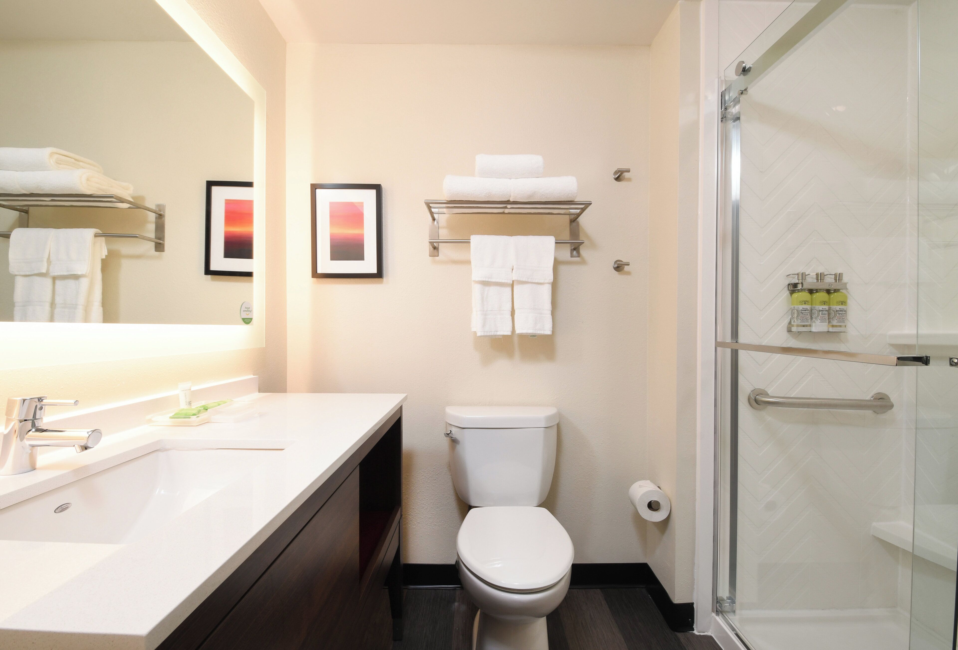 A modern bathroom with a white sink, a toilet, and a glass shower enclosure. There are framed pictures on the wall and a towel rack with folded towels.