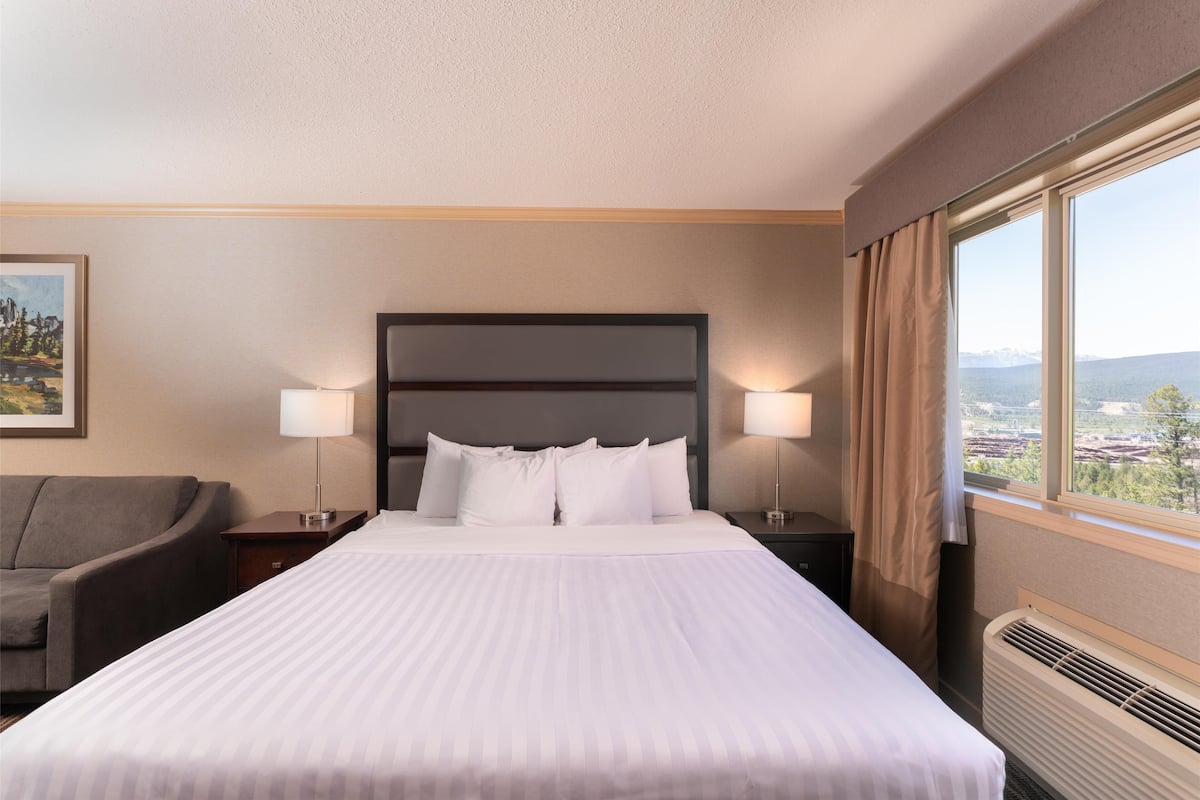 A hotel room with a large bed, two bedside lamps, a nightstand, a chair, and a window with a city view.