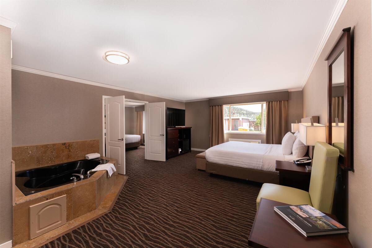 A hotel room with a large bed, a sitting area with a chair, a desk, a bathroom with a bathtub, and a window with curtains.