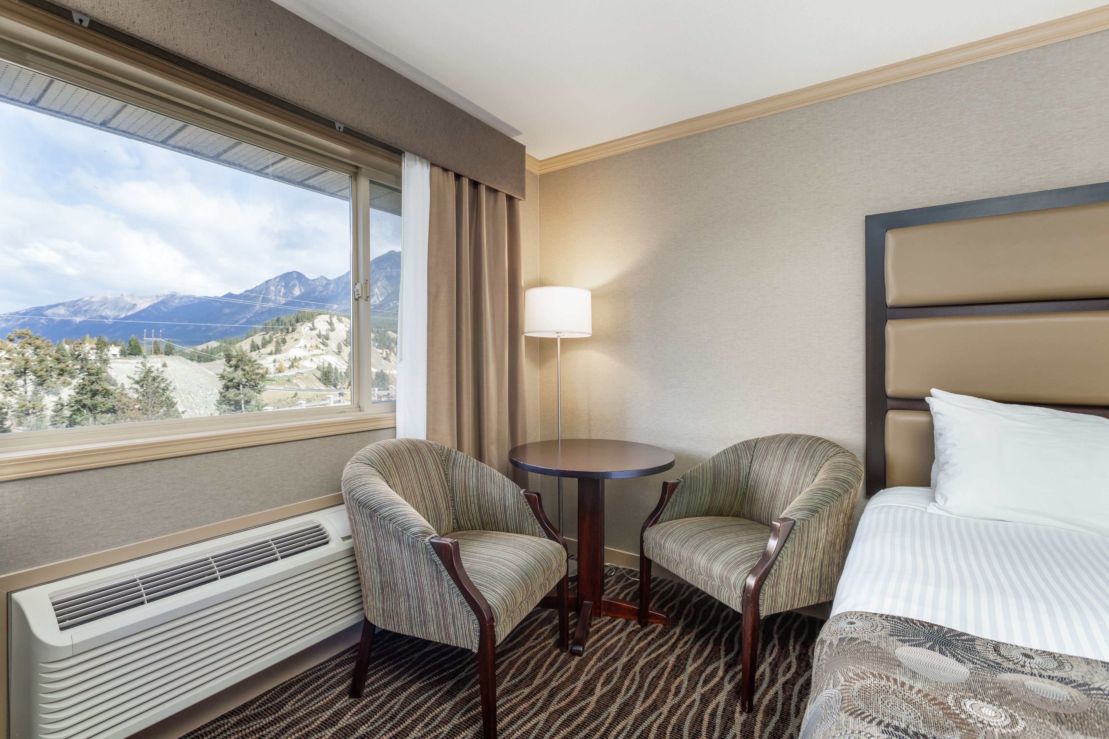 A hotel room with a bed, two armchairs, a small table, a lamp, and a view of mountains through a large window.