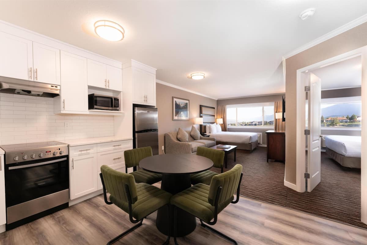 A modern kitchen with white cabinets, a stainless steel refrigerator, and a built-in oven. There is a dining area with a round table and four chairs. The room has a large window offering a view of the outdoors.