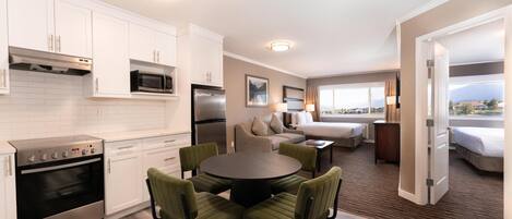 A modern kitchen with white cabinets, a stainless steel refrigerator, and a built-in oven. There is a dining area with a round table and four chairs. The room has a large window offering a view of the outdoors.