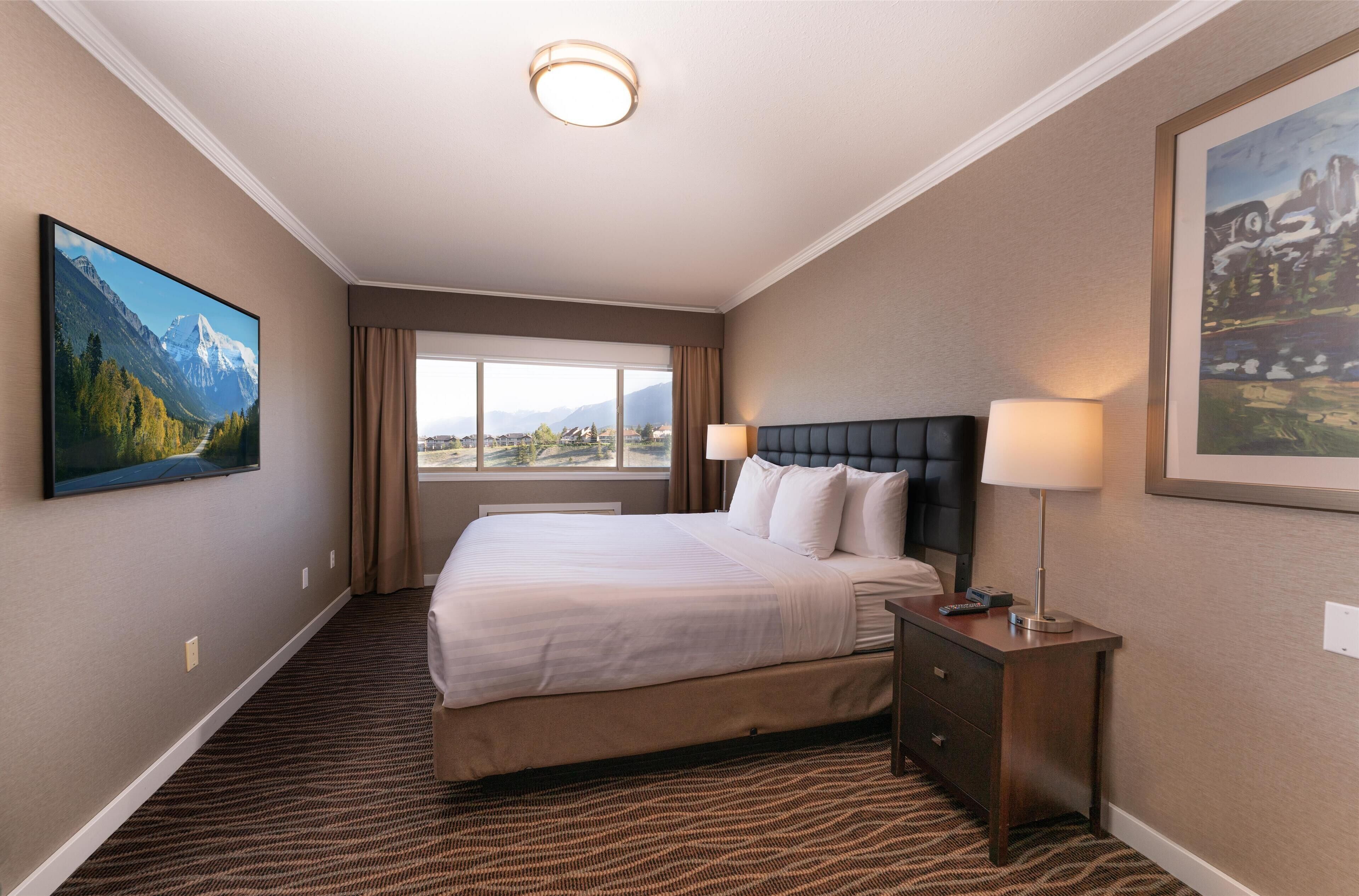 A hotel room with a large bed, a nightstand, a lamp, a TV mounted on the wall, and a window with a view of mountains.
