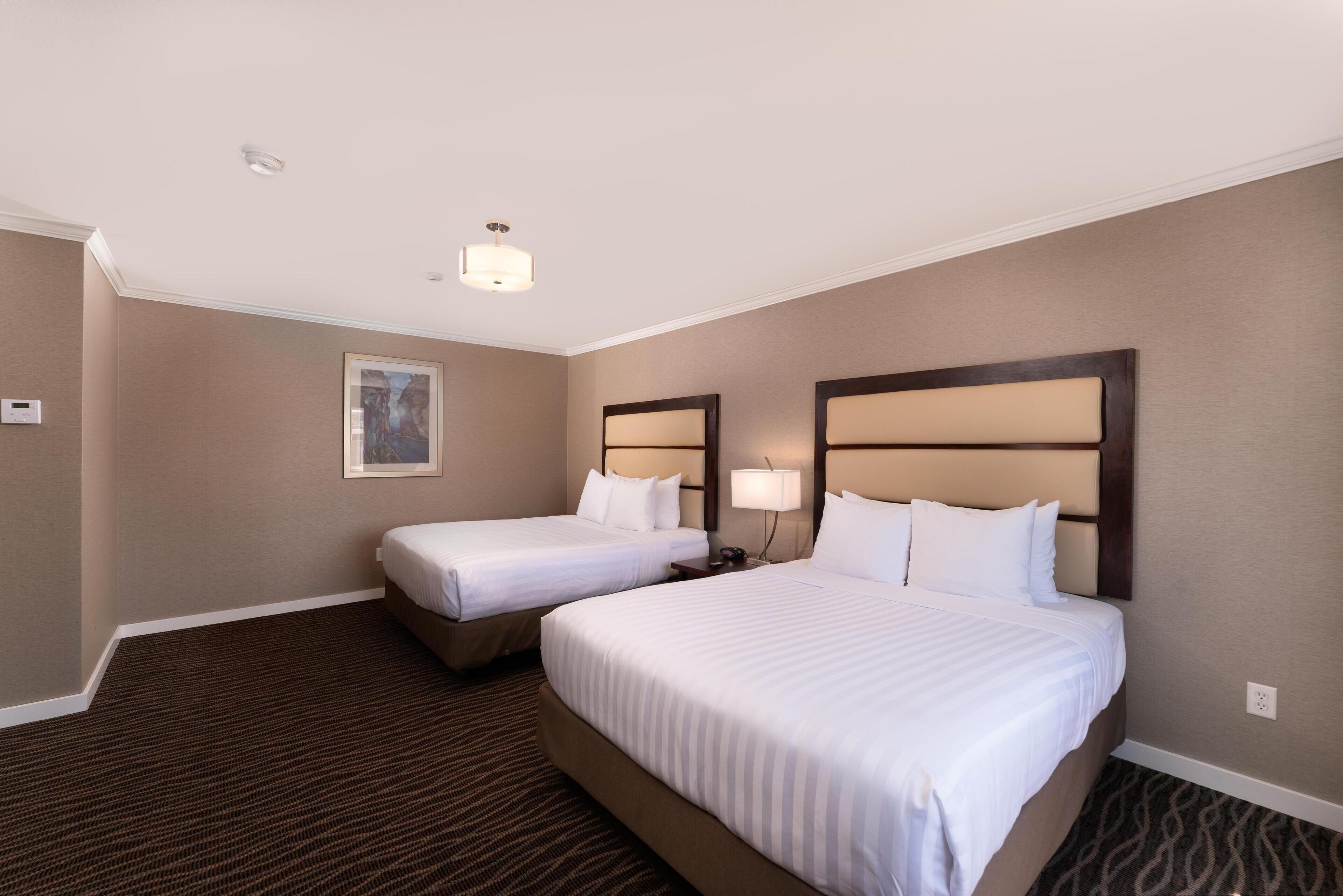 A hotel room with two beds, a carpeted floor, a painting on the wall, and a ceiling light.