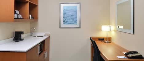 A hotel room with a kitchenette, a desk with a lamp, a telephone, and a framed artwork on the wall.
