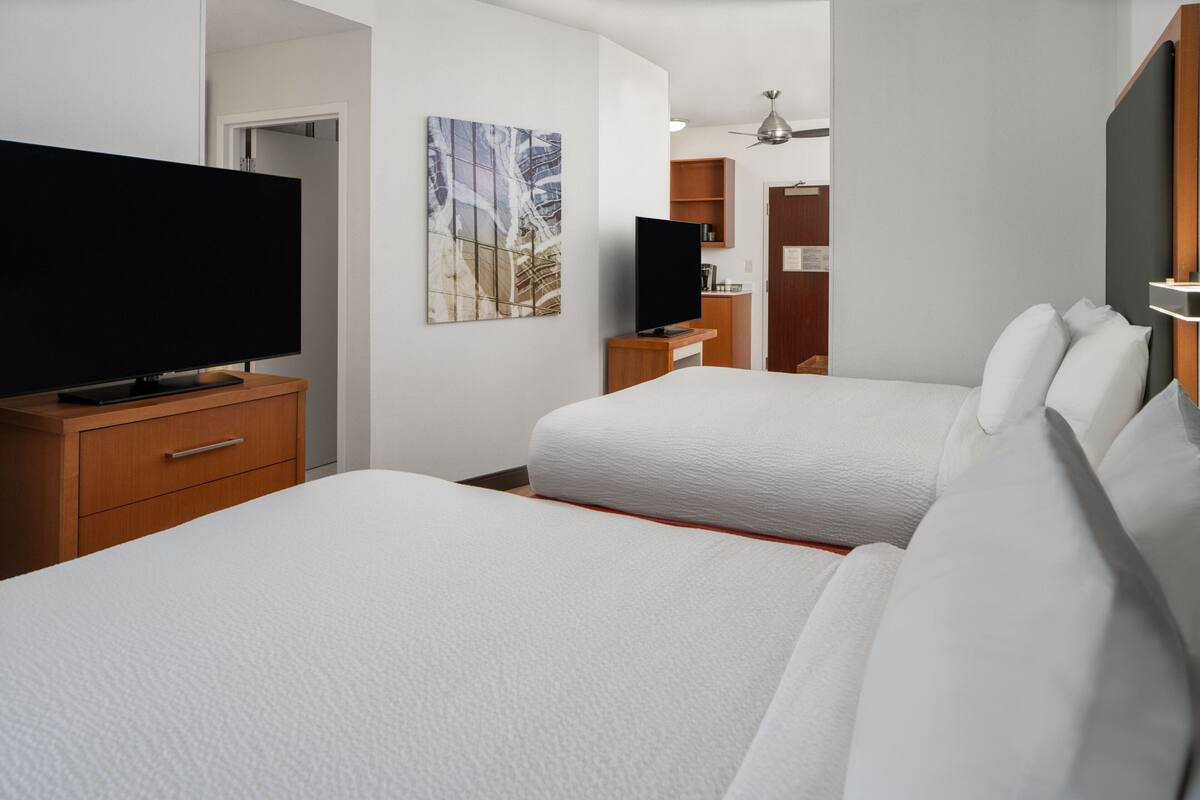 A hotel room with two beds, a TV, a painting, and a small kitchenette.