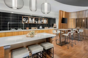 Une cuisine moderne avec un plan de travail blanc, des appareils en acier inoxydable et un pare-feu en bois. On y trouve des tabourets de bar et un téléviseur fixé au mur.