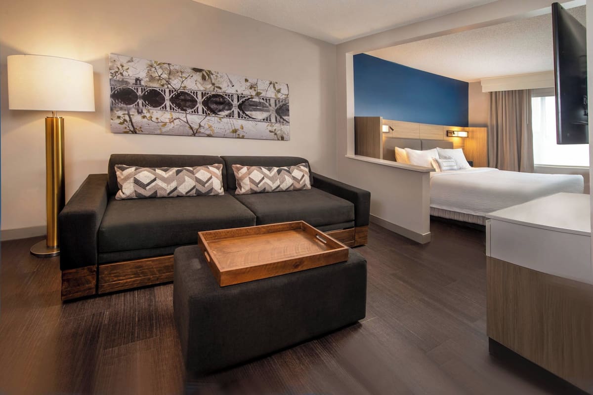 A modern hotel room with a sofa, a wooden tray, a bed, a desk, and a large artwork on the wall.