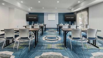 A conference room with a blue patterned carpet, white chairs, and tables. Two flat-screen TVs are mounted on the walls.